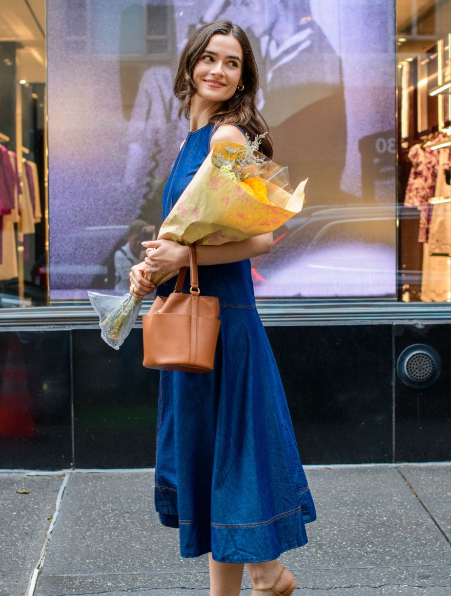 Woman in a blue dress holding flowers and a brown handbag in front of a store window.